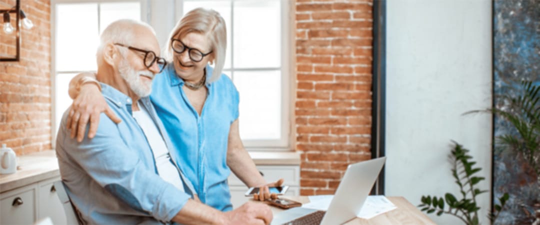 An elderly woman with her arm around an elderly man while looking at a computer