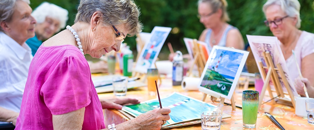 A senior woman outside painting on a canvas with other women.